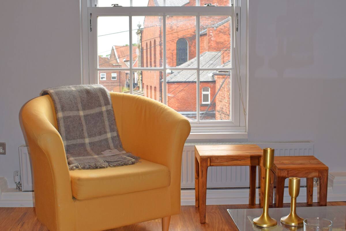 A modern living room with a yellow armchair, a glass coffee table, and a view of a red brick building through a large window.