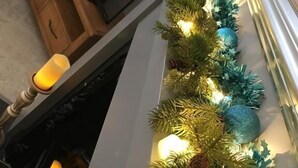 A fireplace with a garland of green foliage and blue ornaments, a wooden cabinet, and a lit candle. - Ourhaus (Beverley)