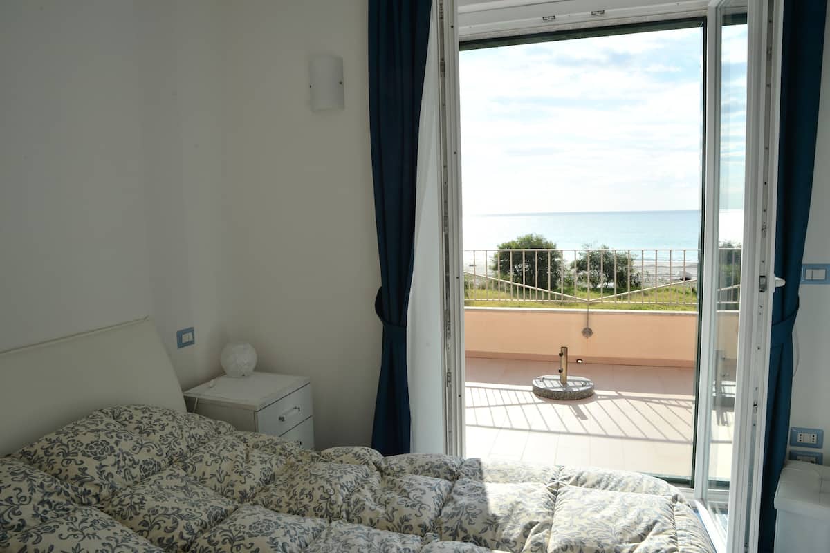 Un dormitorio con cama, mesita de noche, lámpara y vista al mar a través de una puerta de balcón.