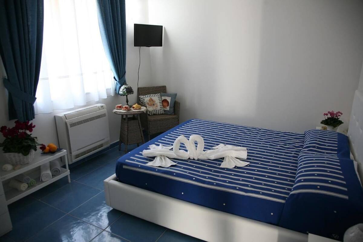 Una habitación de hotel con una cama, una mesita con lámpara, una silla, un estante con toallas y una ventana con cortinas.
