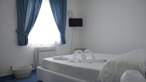 Habitación de hotel con cama, televisión montada en la pared, ventana con cortinas azules, radiador y una pequeña cesta.