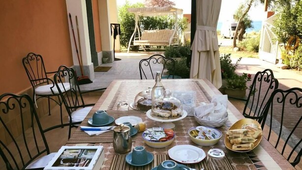 Un área de comedor al aire libre con una mesa dispuesta para una comida, rodeada de sillas, y vistas al jardín y al mar al fondo.