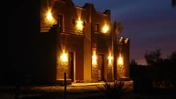 A building with multiple illuminated windows and doors, surrounded by a dry landscape.