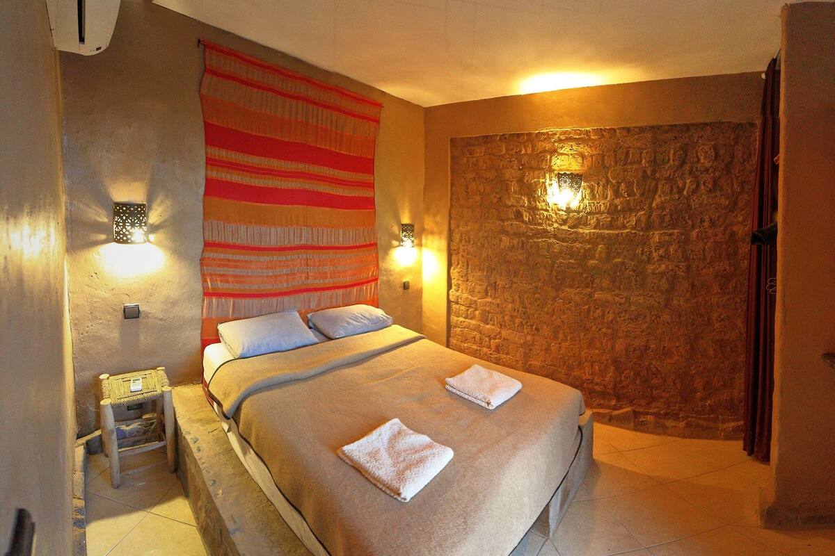 Un dormitorio con cama, mesita de noche, lámpara de pared y pared de piedra texturizada.