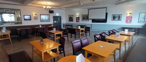 A restaurant with wooden tables and chairs, a bar area, and a projection screen on the wall.