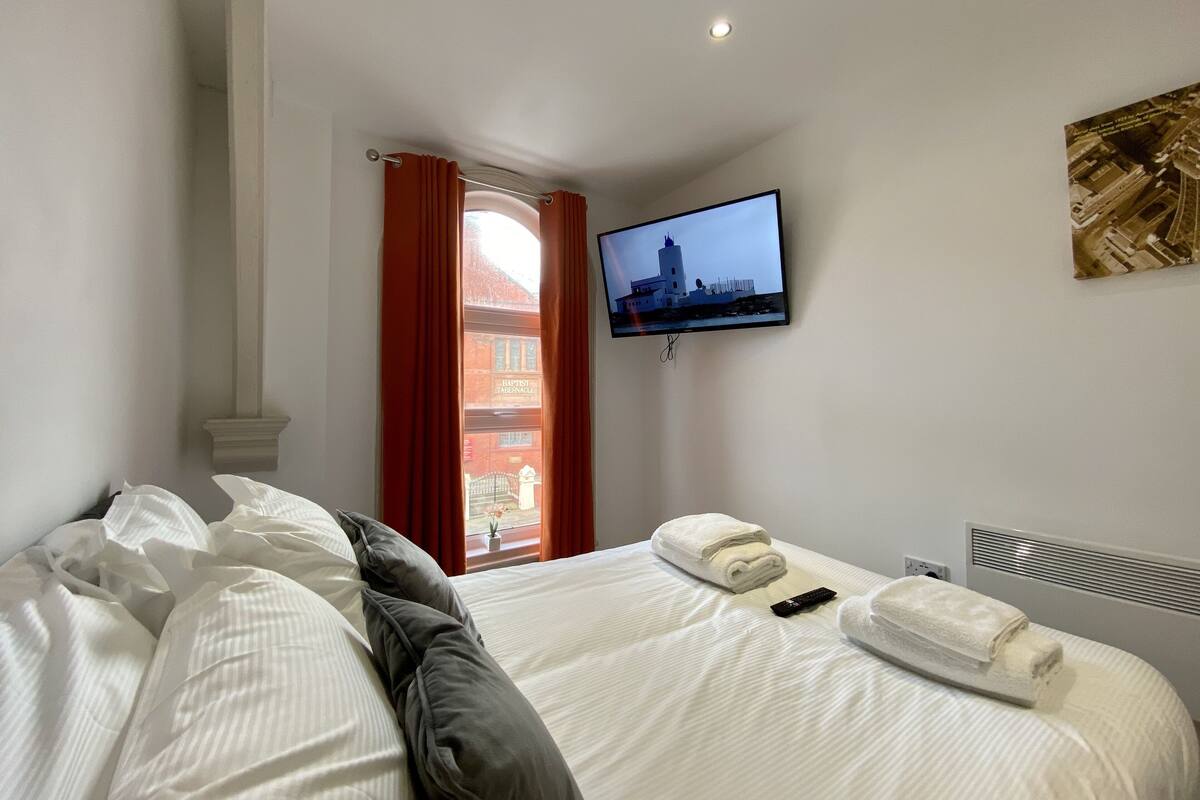 A hotel room with a bed, a television mounted on the wall, and a window with red curtains.