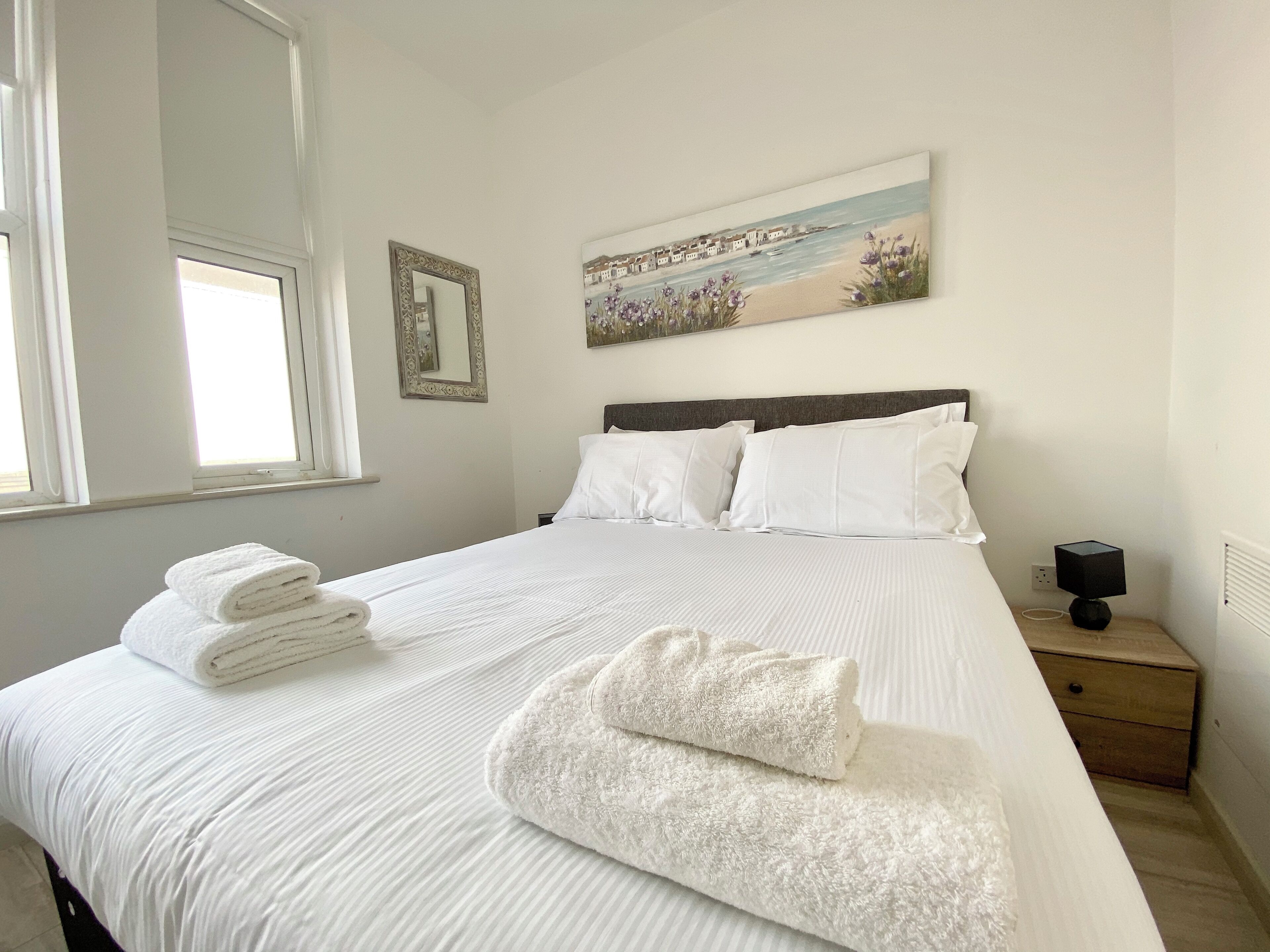 A neatly made bed with white linens and towels, a painting of a beach scene above it, and a small bedside table with a lamp.