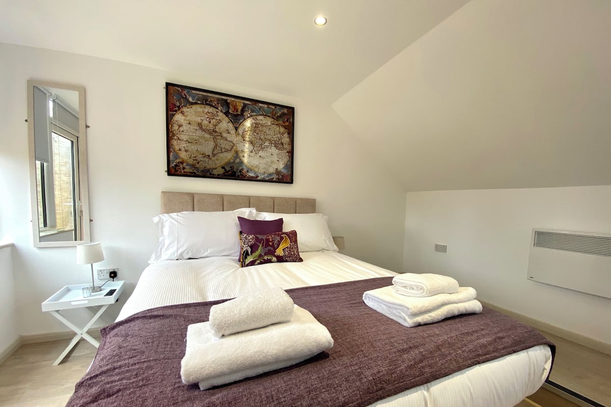 A neatly made bed with white and purple bedding, a world map artwork above it, and a window on the left.