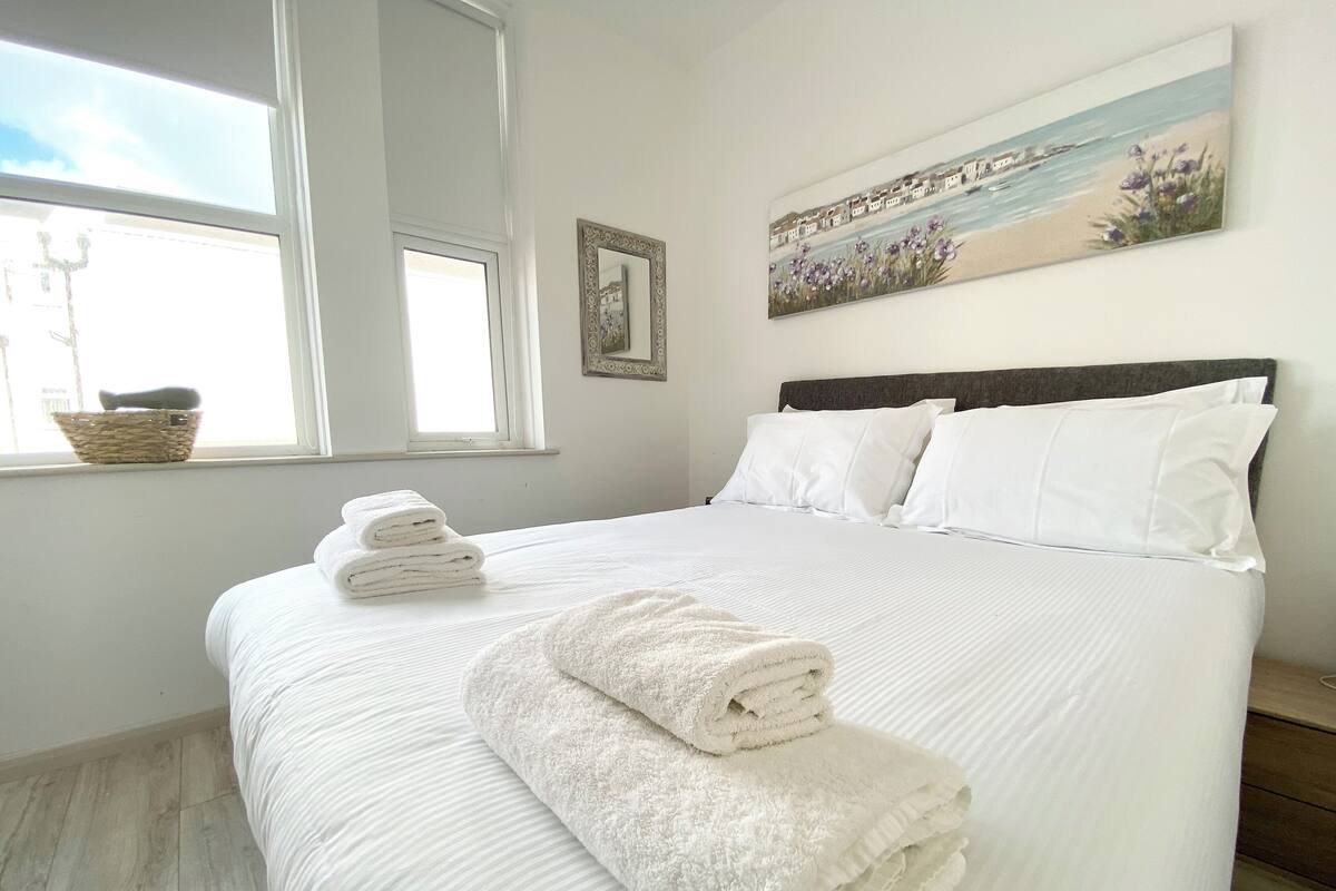 A neatly made bed with white linens and a fluffy white towel on the footboard. There is a window with a basket on the sill and a painting above the bed.
