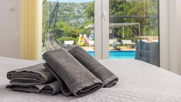 A neatly folded gray towel on a bed with a white quilted cover, a large window with a view of a swimming pool and outdoor furniture, and a yellow vertical element on the wall.