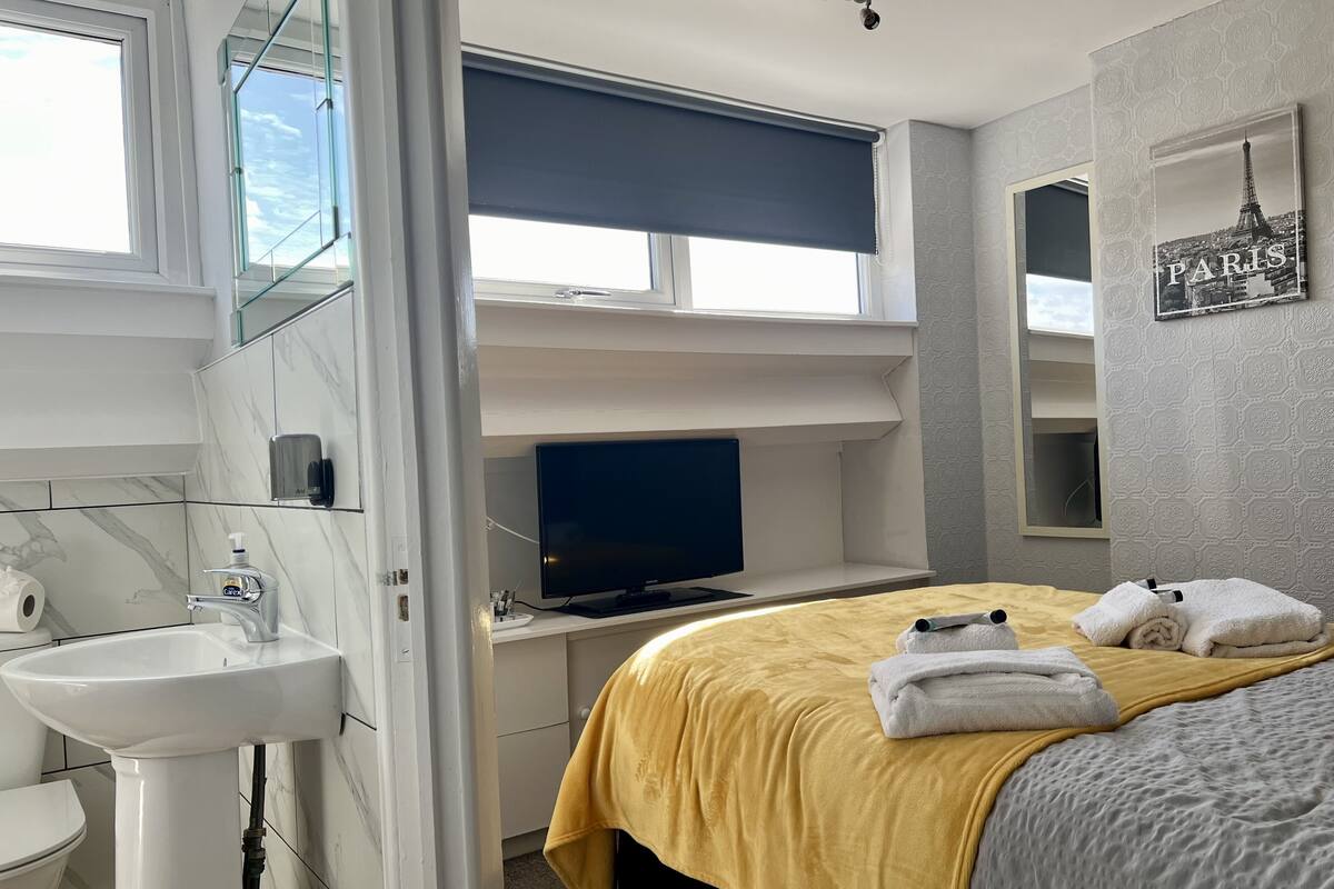 Une chambre d’hôtel moderne équipée d’un lit, d’un bureau avec un ordinateur, d’une salle de bain avec un lavabo et des toilettes, et d’une fenêtre donnant sur le ciel.