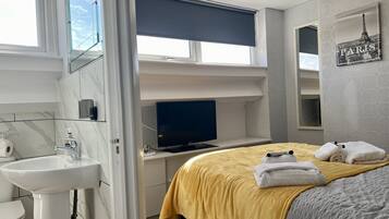 Quarto de hotel moderno com cama, escrivaninha com computador, banheiro com pia e vaso sanitário, e janela com vista para o céu.