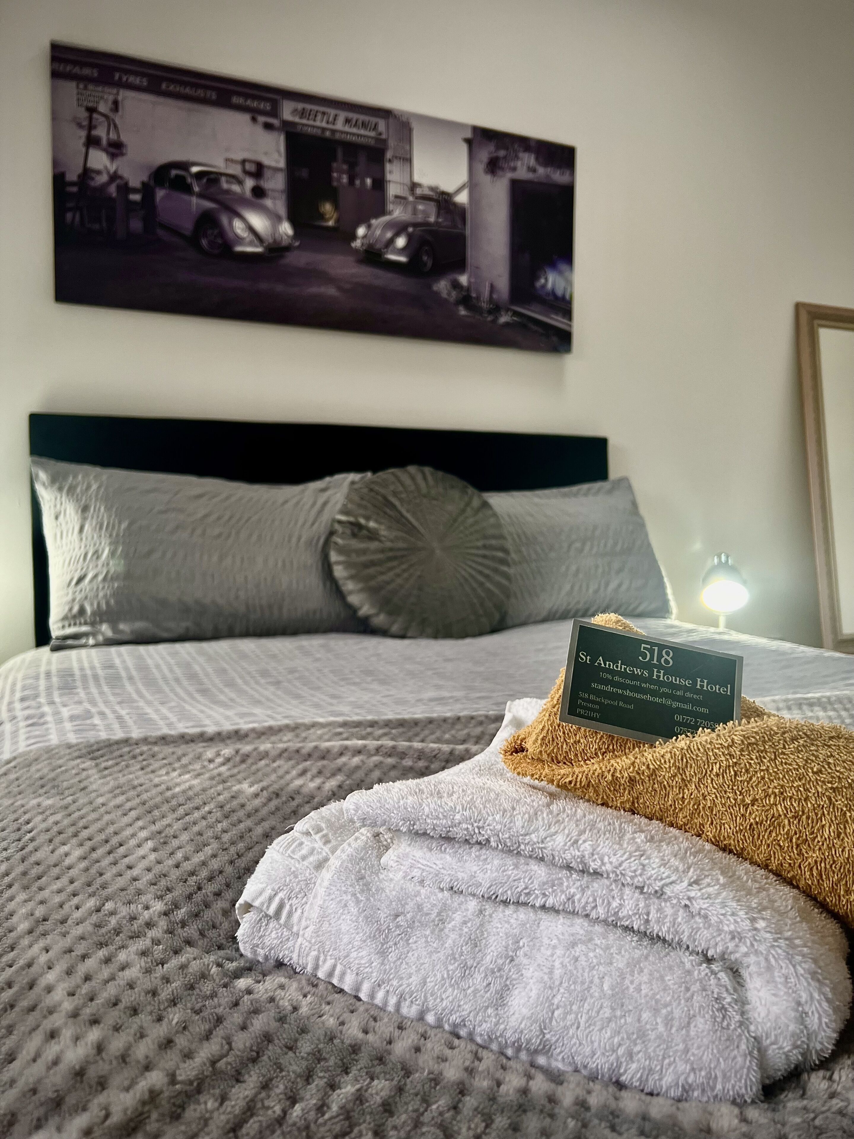 A hotel room with a bed, pillows, a blanket, a sign with the hotel name and address, and a wall-mounted picture of a car and garage.