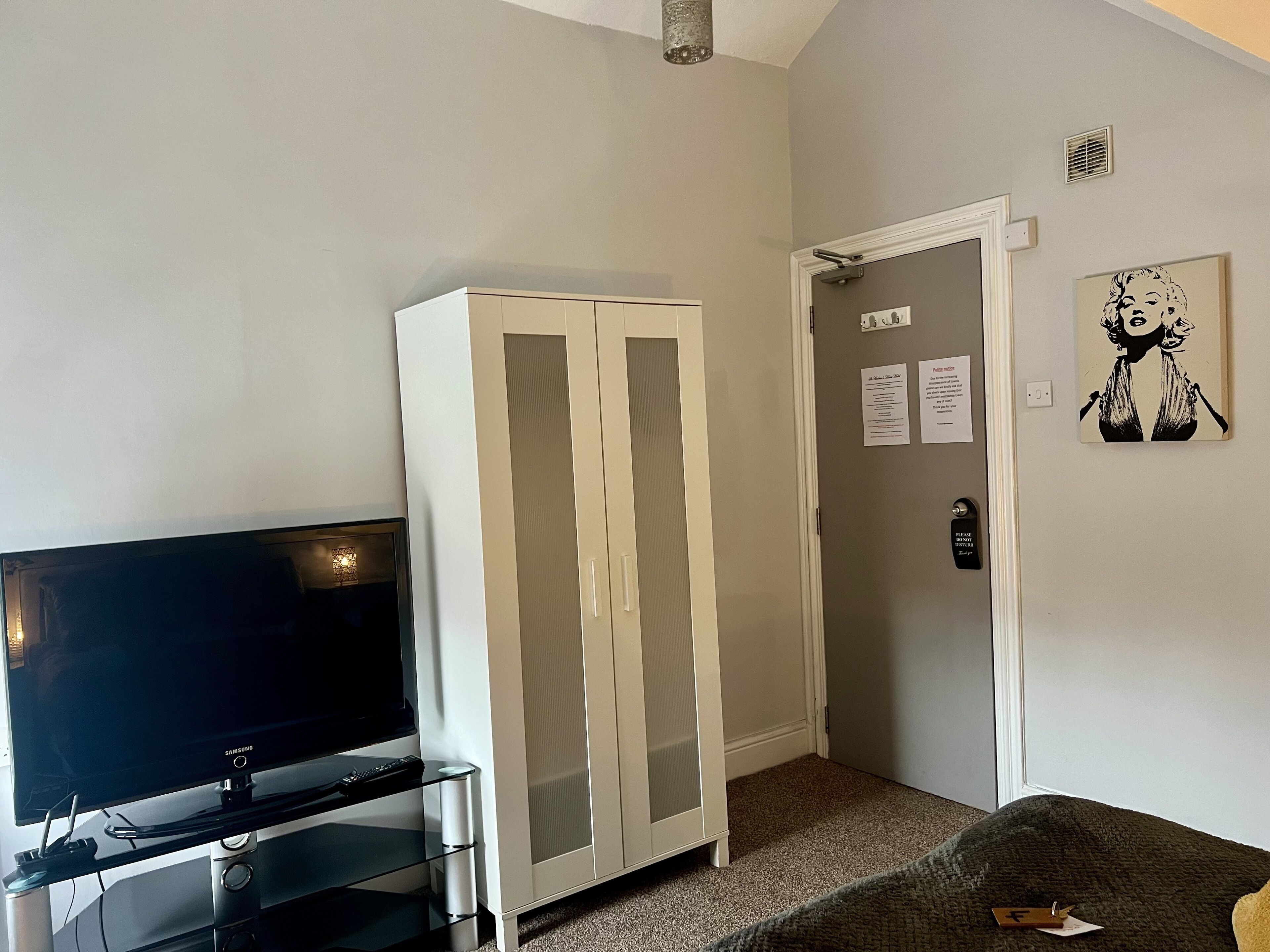 Una habitación con una televisión de pantalla plana sobre un soporte, un armario blanco, una puerta con un letrero y un cuadro en la pared.