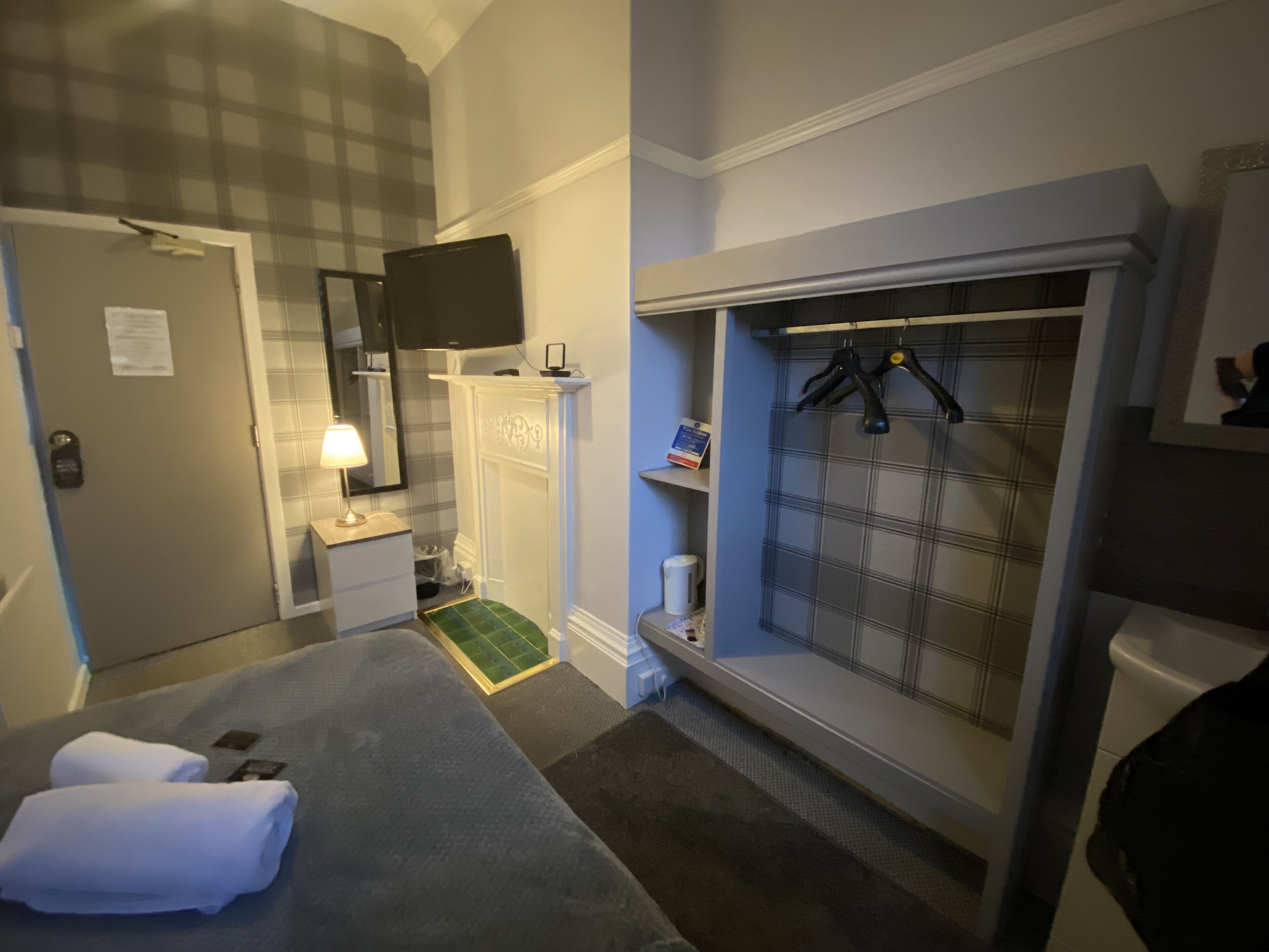 A hotel room with a bed, a desk, a mirror, a television, a wardrobe, and a small shelf with toiletries.