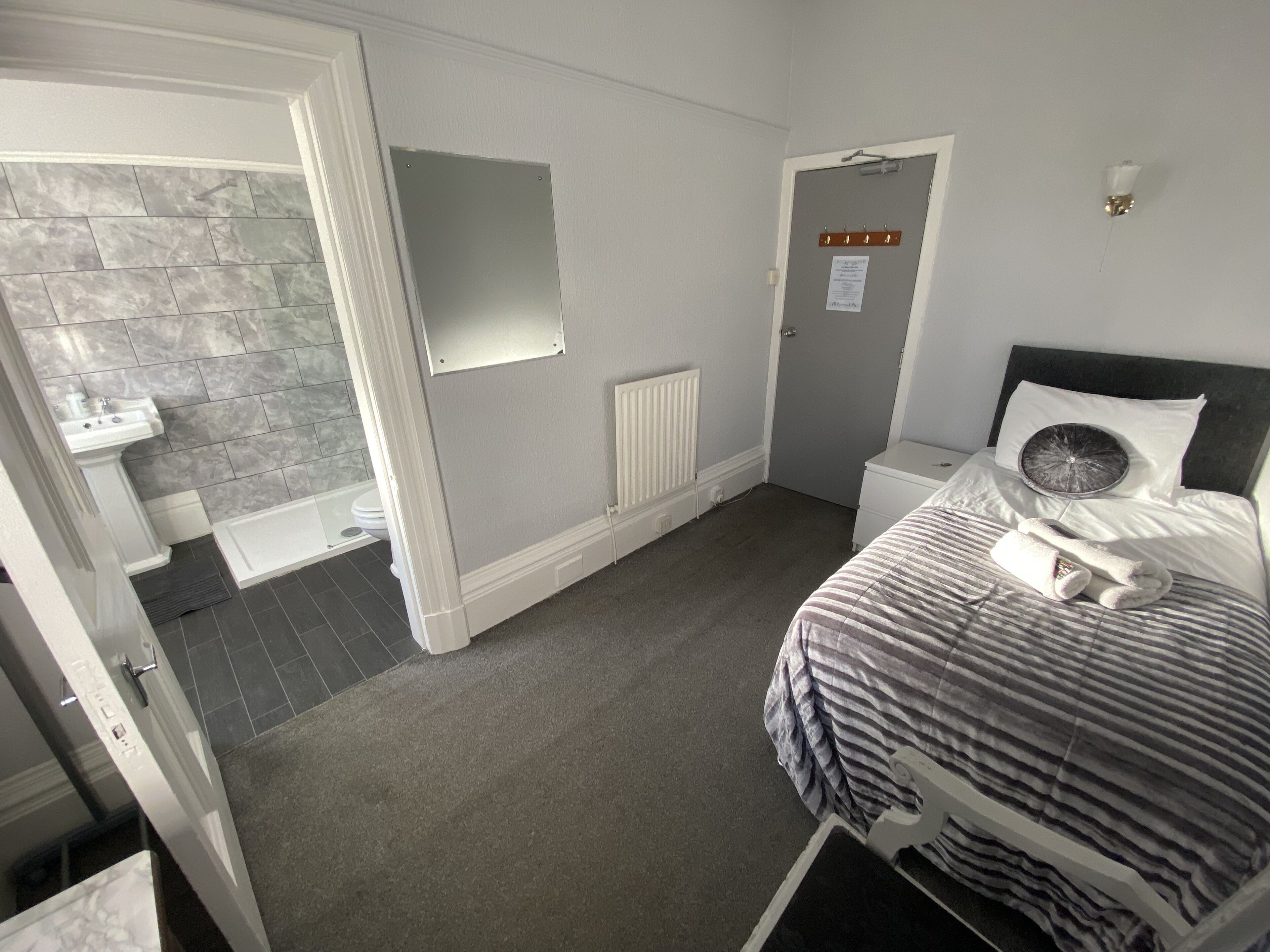 Habitación de hotel con cama, mesita de noche, tocador pequeño, espejo, radiador y baño con lavabo e inodoro.