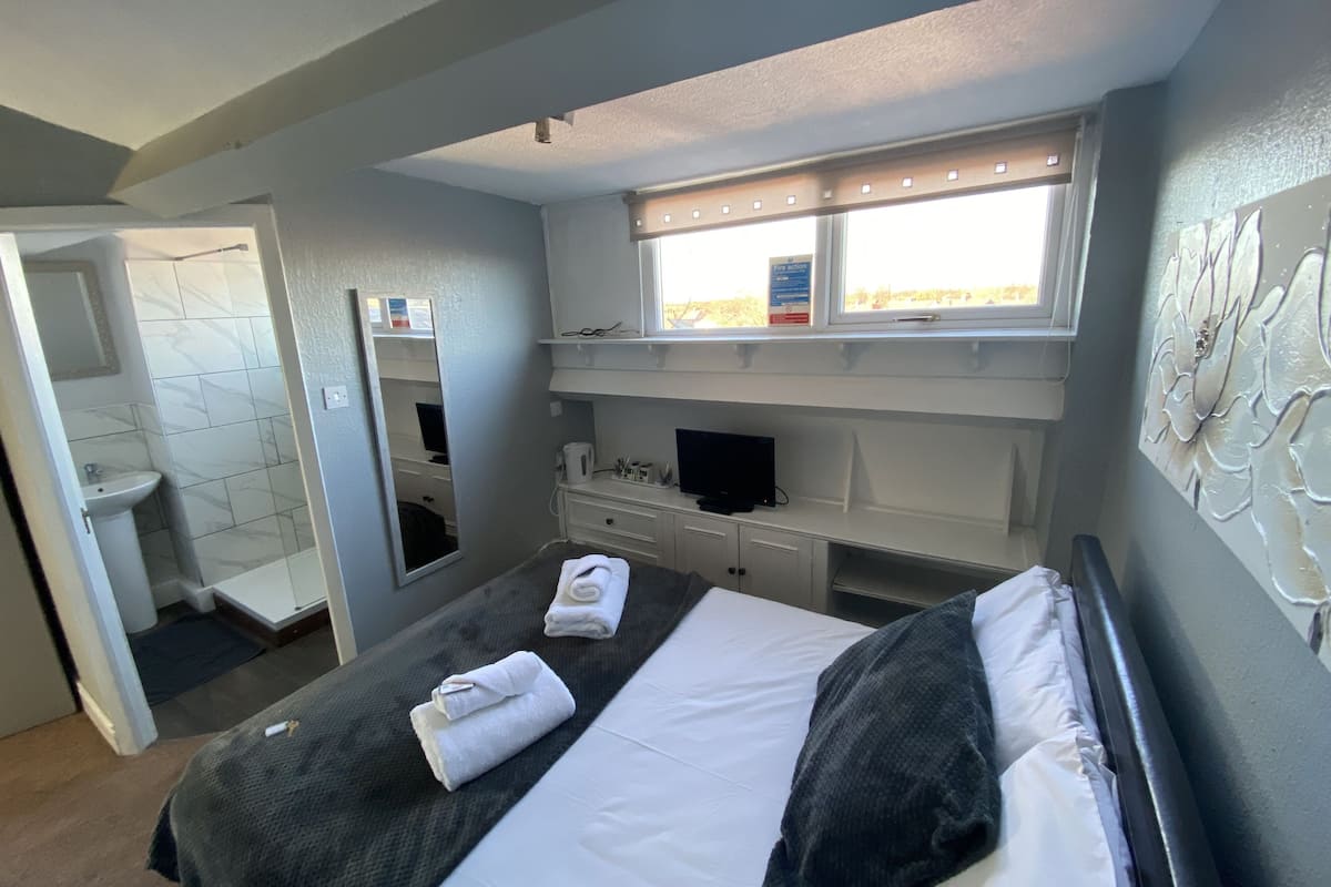 Une chambre d’hôtel moderne équipée d’un lit, d’une télévision, d’une fenêtre avec stores, d’une salle de bain avec un lavabo et un miroir, et d’un mur agrémenté d’un panneau décoratif.