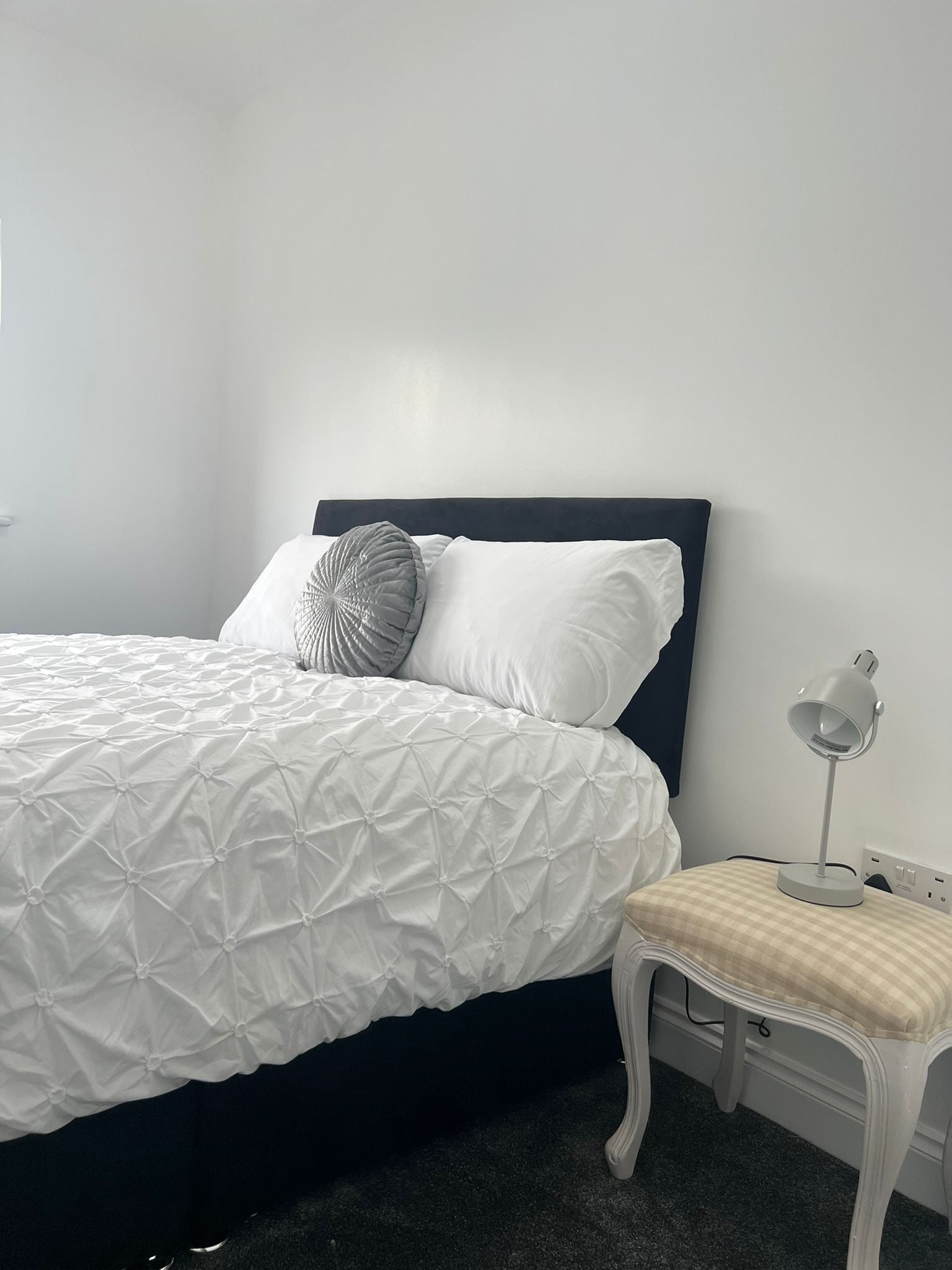 A neatly made bed with a white duvet and two pillows, a bedside table with a lamp, and a chair with a checkered cushion.