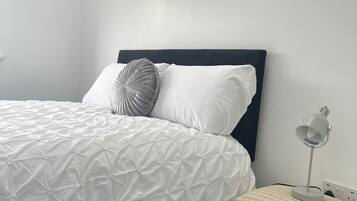 A neatly made bed with a white duvet and two pillows, a bedside table with a lamp, and a chair with a checkered cushion.