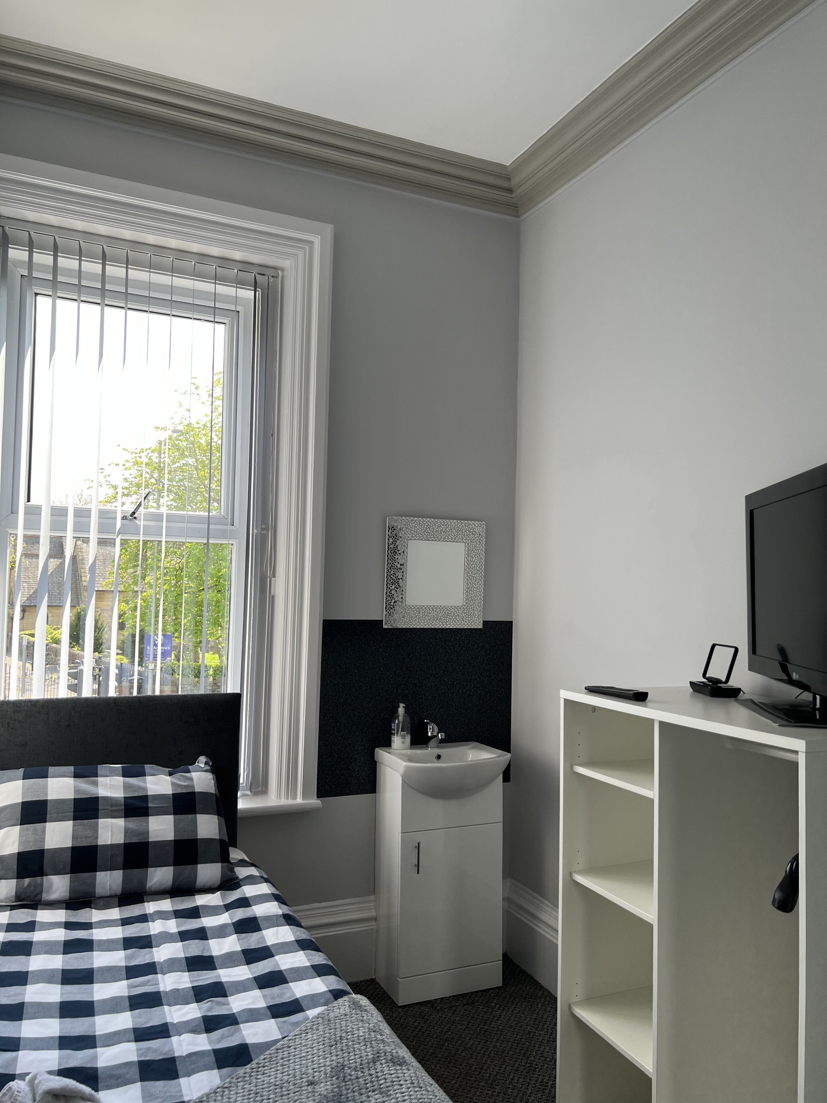 Un dormitorio con una cama, una ventana, una mesita de noche con espejo, un televisor y un armario blanco.