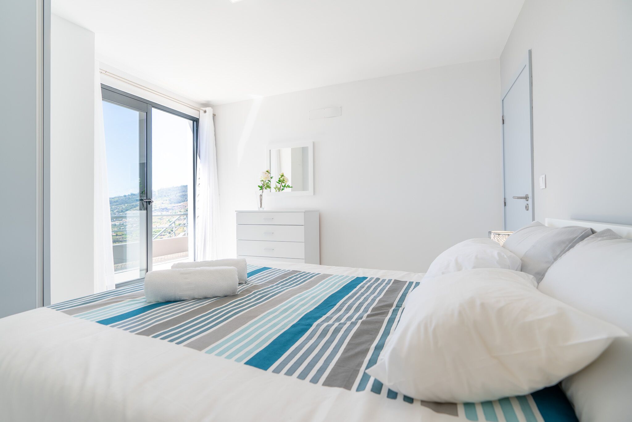 Une chambre d’hôtel moderne avec un grand lit, un balcon offrant une vue sur la ville, une commode blanche et une petite plante posée sur la commode.
