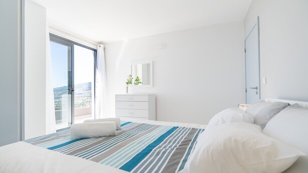 Ett modernt hotellrum med en stor säng, en balkong med utsikt över staden, en vit byrå och en liten växt placerad på byrån.