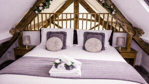 Ein Schlafzimmer mit hoher Decke, Holzbalken, einem Bett mit zwei Kissen, einem Nachttisch mit Lampe und einem Fenster.