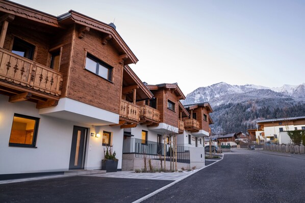 A row of wooden buildings with balconies, a paved street, and snow-capped mountains in the background.