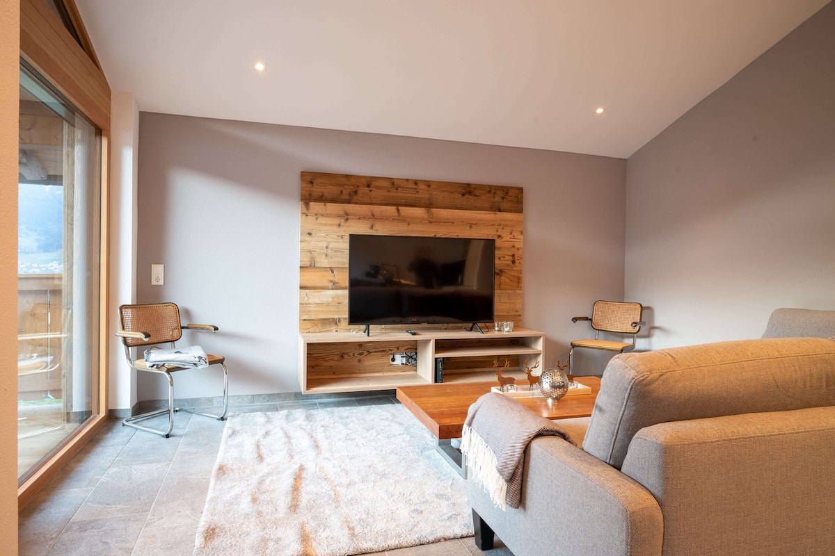 Un salon moderne avec un grand téléviseur à écran plat fixé à un mur en bois, un canapé confortable, une table basse en bois et une porte en verre donnant sur un balcon.