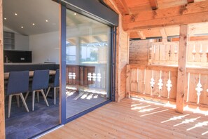 A wooden interior with a glass door leading to an outdoor area, a dining table with chairs, and a kitchen visible through the glass.
