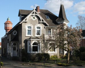 A two-story house with a steeply pitched roof, a prominent tower, and a symmetrical facade. - Alleehotel Eschen (Aurich)