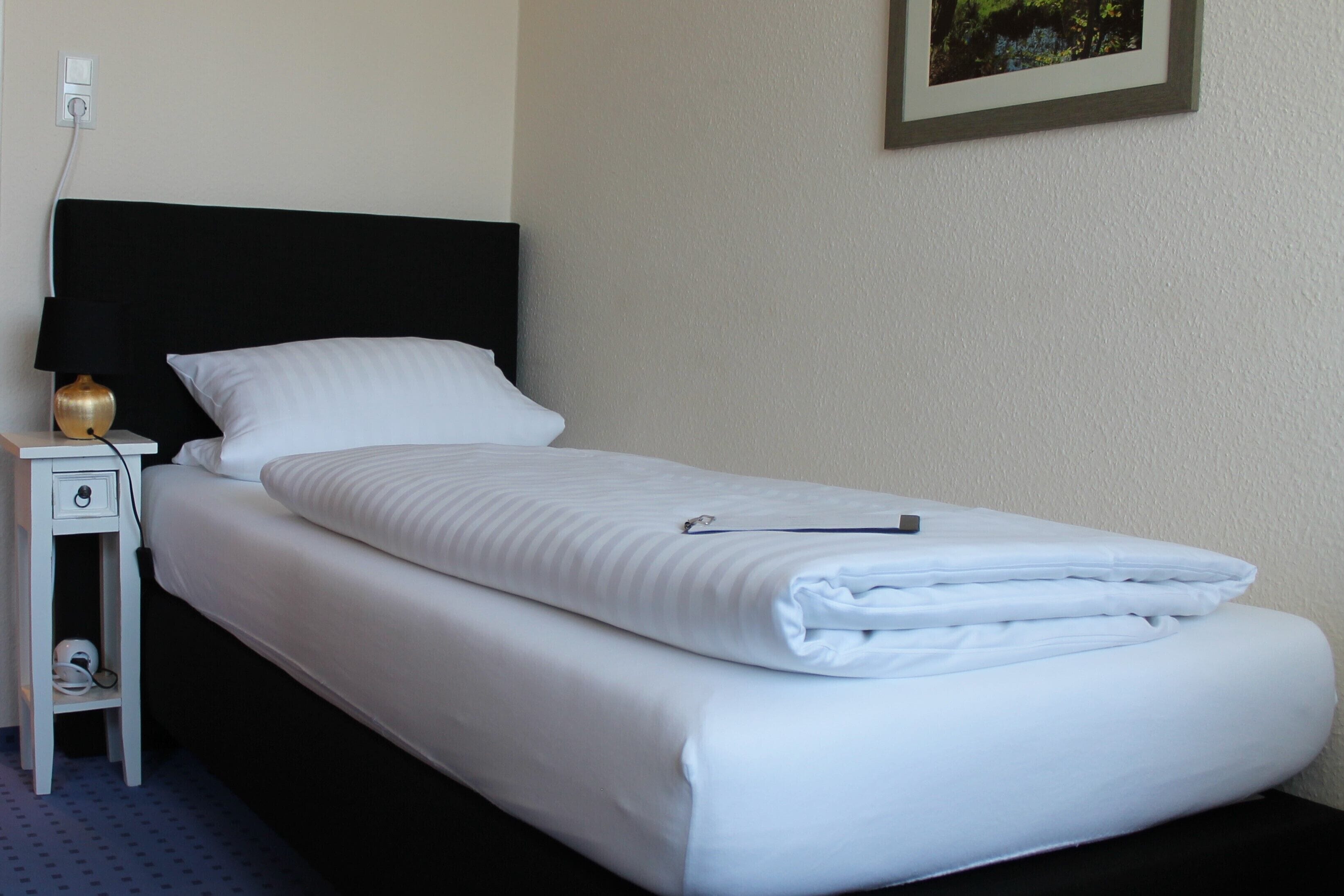 A neatly made bed with white bedding, a black headboard, a small bedside table with a lamp, and a power outlet on the wall.