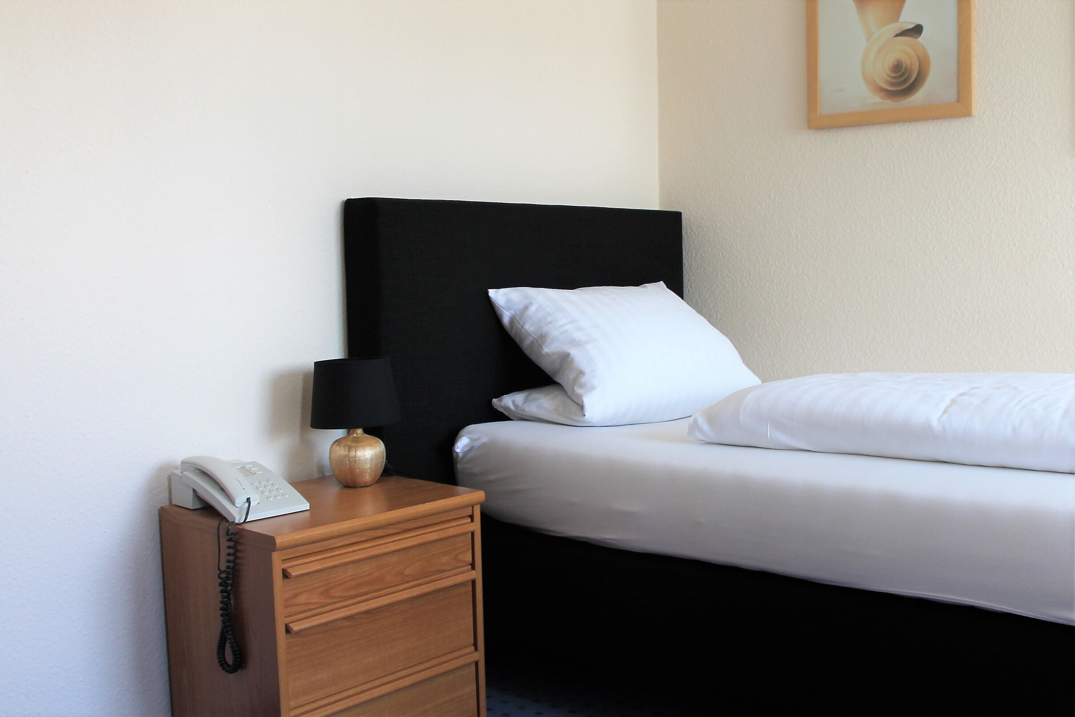Hotellihuone, jossa on sänky, puinen yöpöytä, jossa on puhelin ja lamppu, sekä seinällä oleva kehystetty kuva.