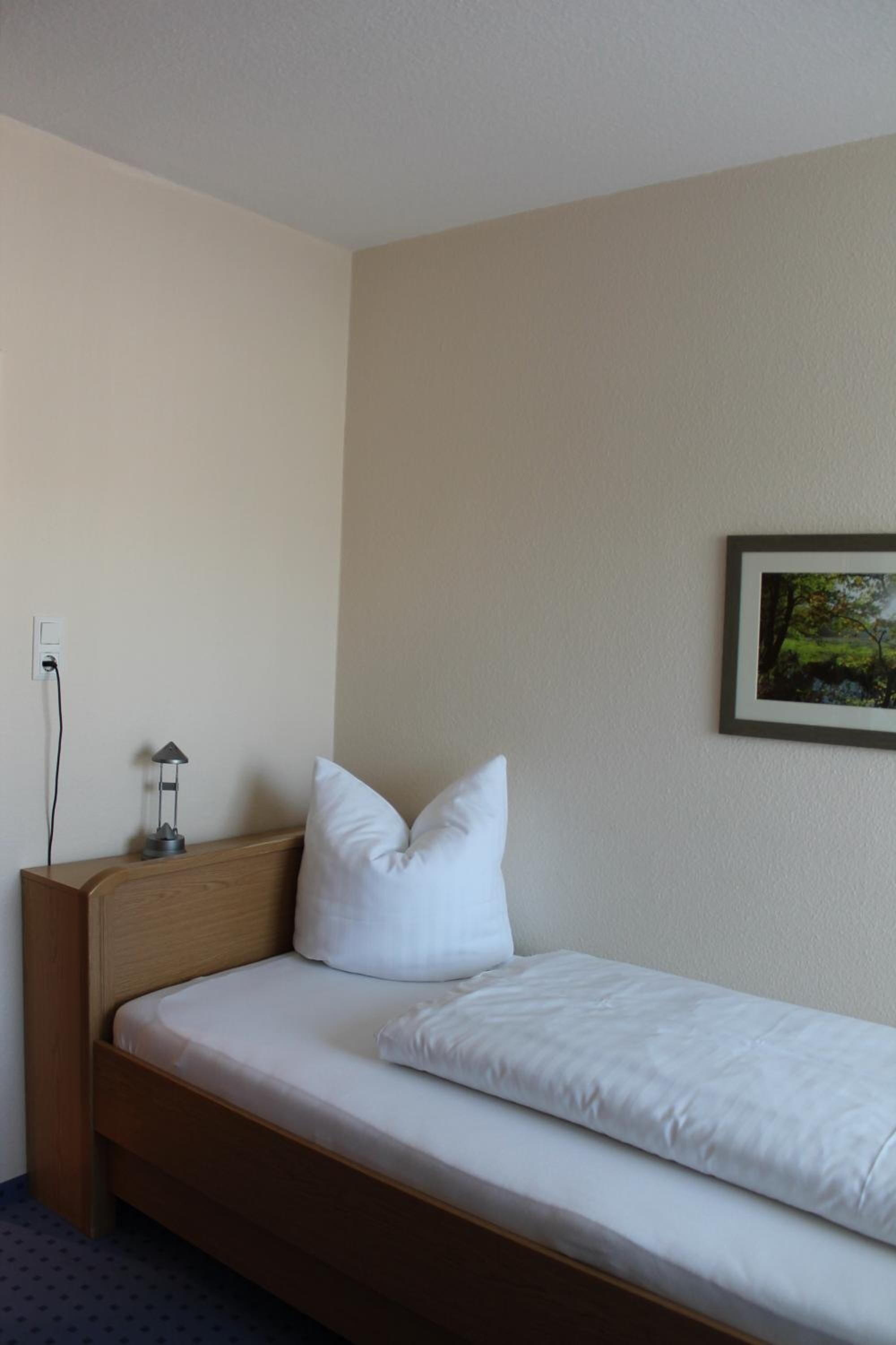 Una cama individual con ropa de cama blanca, cabecera de madera y mesita de noche con lámpara.