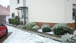 Eine Wohnstraße, auf der linken Seite ist ein rotes Auto geparkt. Schnee bedeckt den Boden und die Büsche entlang des Bürgersteigs.