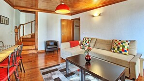 A modern living room with a wooden ceiling, a sofa with colorful cushions, a dining area with chairs, and a staircase.