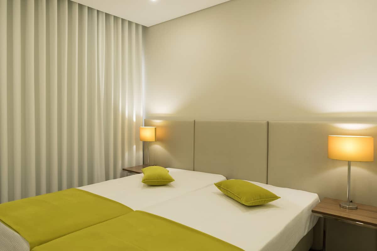 A hotel room with two beds, each with a green pillow, a bedside table with a lamp, and a window with vertical blinds.