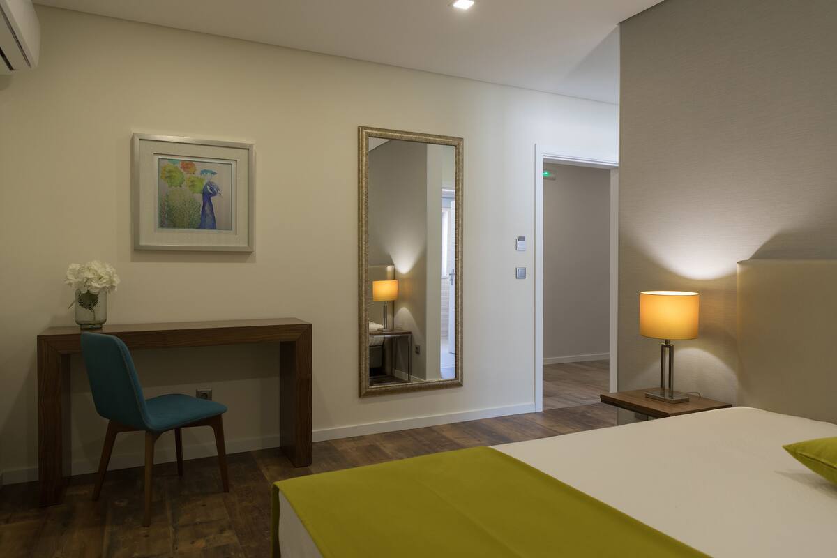 A hotel room with a bed, a desk with a chair, a mirror, a lamp, and a painting on the wall.