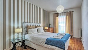 Une chambre moderne avec un grand lit, une table en verre, une table de chevet, un luminaire suspendu et une fenêtre avec des rideaux.
