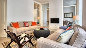 A living room with a sofa, armchair, and coffee table, a TV mounted on the wall, and a dining area visible through an open door. - MyPlace - Lisbon - Luxury Trindade (Lisboa)