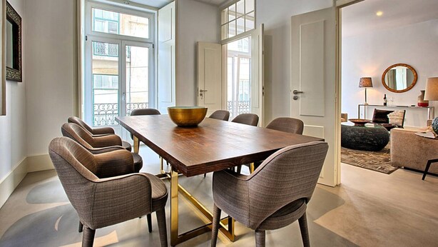 A dining area with a wooden table and six upholstered chairs, large windows, and a view of the city. - MyPlace - Lisbon - Luxury Trindade (Lisboa)