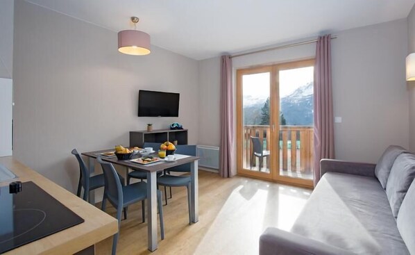 A modern hotel room with a dining area, a sofa, a TV, and a balcony with a mountain view.