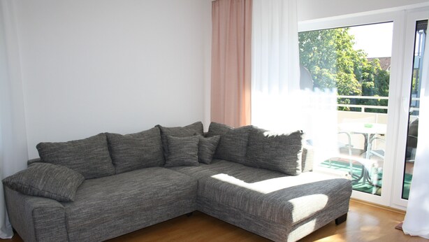 A grey sectional sofa with multiple cushions in a room with wooden flooring and a large sliding glass door leading to an outdoor area. - Apartment Bad Rappenau (Bad Rappenau)