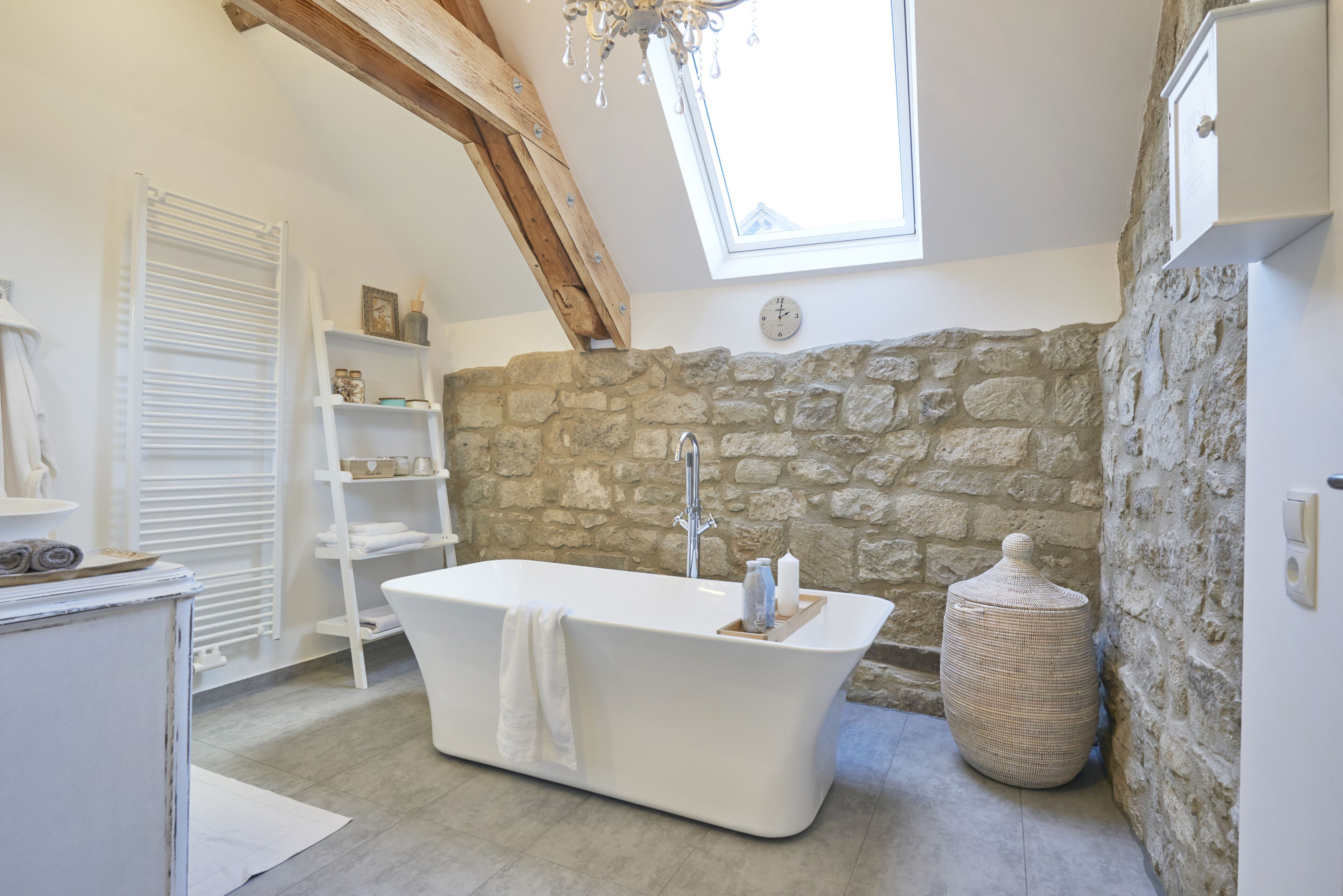 A bathroom with a freestanding bathtub, a stone wall, a wooden beam ceiling, and a skylight.