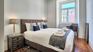 A neatly arranged bedroom with a large bed, bedside lamps, a dresser, and a view of buildings through a window.
