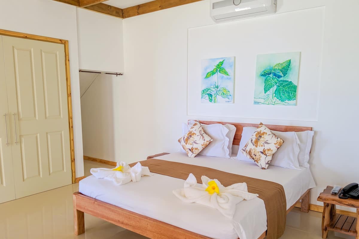 A neatly made bed with floral pillows, a wooden bedside table with a phone, and two framed botanical paintings on the wall.