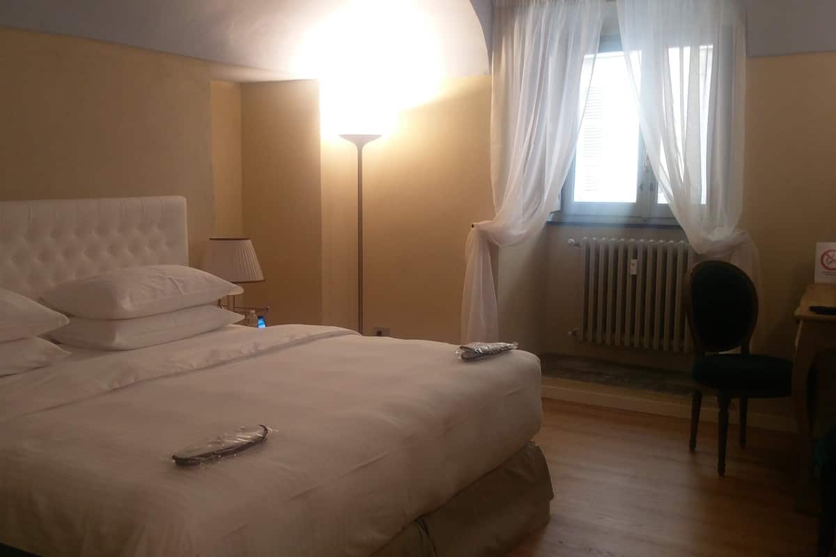 A neatly made bed with pillows and a remote control, a bedside lamp, a chair, and a window with sheer curtains.