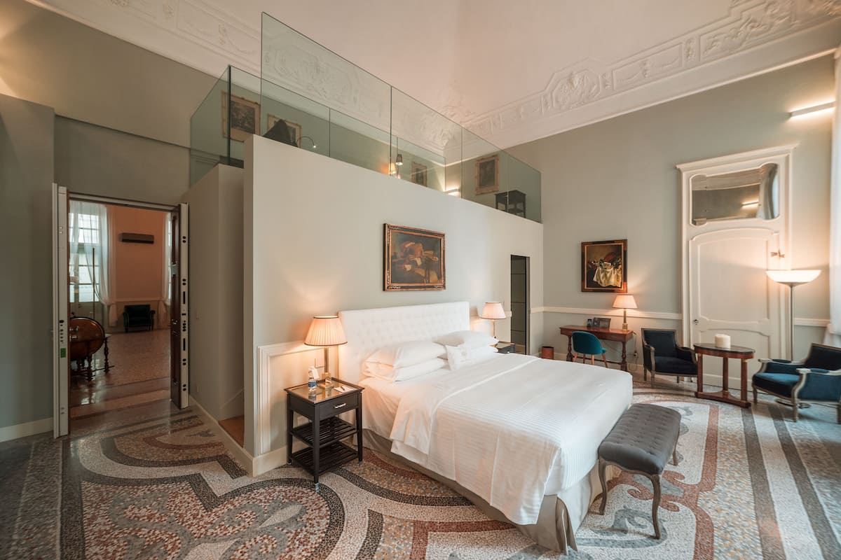 A hotel room with a large bed, a glass balcony, a desk with a chair, and a small table with a lamp.