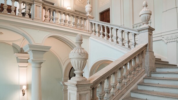 A grand staircase with ornate balustrades and a chandelier, leading to an upper level with decorative moldings and a door. - Royal Palace Luxury Suites & Hotel (Turin)