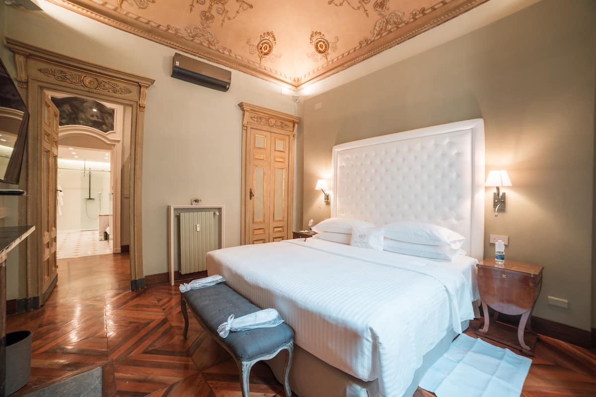 A hotel room with a large bed, a bench, a nightstand, a mirror, a door, and a ceiling with decorative molding.